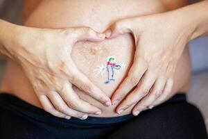 Painted boy on his belly of a pregnant woman. She creates a heart around the drawing with her hands. photo