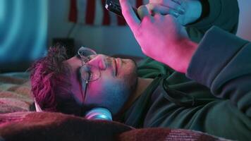 joven pelinegro chico en auriculares y lentes usos su teléfono mientras acostado en el cama. medio disparo. 4k video