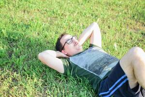 joven hombre con lentes y verde camiseta haciendo Abdominales y hacer ejercicio en parque foto