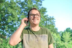 joven hombre con los anteojos y verde camiseta hablando en célula teléfono a el parque foto