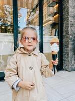 buda, montenegro - 18 diciembre 2023. pequeño niña en Gafas de sol posando con un grande hielo crema cono en su mano cerca el vaso puerta de el edificio foto