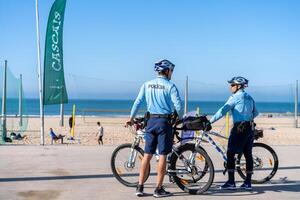 Portugal, Carcavelos April 2022 Two policeman patrolling seaside promenade on bicycles. People are sunburning on the city public beach on the Atlantic shore. photo