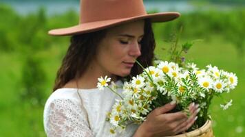 manzanilla mujer. contento mujer en un blanco vestir y marrón sombrero sostiene un ramo de flores de margaritas en su manos y huele ellos video