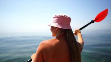 een vrouw in een roze hoed peddels een kajak in de oceaan. de tafereel is vredig en ontspannend, met de vrouw genieten van de water en de zon. video