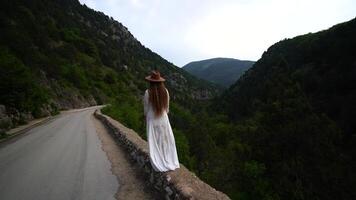 Female traveler in brown hat and white dress looking at amazing mountains and forest, wanderlust travel concept, atmospheric epic moment video