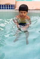 Happy Indian boy swimming in a pool, Kid wearing swimming costume along with air tube during hot summer vacations, Children boy in big swimming pool. photo