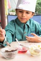 Cute Indian chef boy preparing sundae dish as a part of non fire cooking which includes vanilla ice cream, brownie, coco powder, freshly chopped fruits and strawberry syrup. Little kid preparing food photo