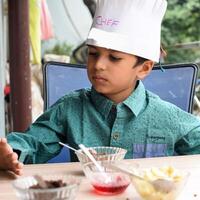 Cute Indian chef boy preparing sundae dish as a part of non fire cooking which includes vanilla ice cream, brownie, coco powder, freshly chopped fruits and strawberry syrup. Little kid preparing food photo