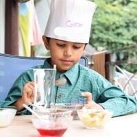 Cute Indian chef boy preparing sundae dish as a part of non fire cooking which includes vanilla ice cream, brownie, coco powder, freshly chopped fruits and strawberry syrup. Little kid preparing food photo