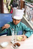 Cute Indian chef boy preparing sundae dish as a part of non fire cooking which includes vanilla ice cream, brownie, coco powder, freshly chopped fruits and strawberry syrup. Little kid preparing food photo