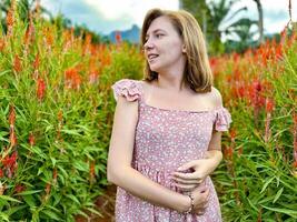 Smiling young woman in a floral summer dress standing in a vibrant red flower field with a scenic mountain backdrop, capturing the essence of summer photo