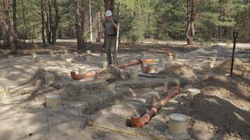 une ouvrier, habillé dans travail tenue et équipé avec sécurité engrenage, est dans le processus de pose grand souterrain tuyaux parmi le partiellement enterré béton fondations dans une boisé construction placer. video