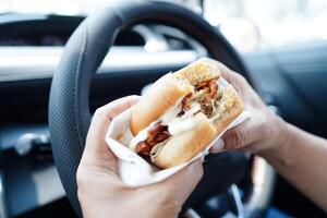 asiático mujer conductor sostener y comer hamburguesa en auto, peligroso y riesgo un accidente. foto