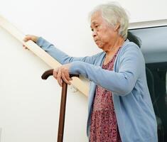 Asian senior woman use walking stick with caregiver help support walking down the stairs prevent accident, slip and fall at home. photo