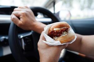 asiático mujer conductor sostener y comer hamburguesa en auto, peligroso y riesgo un accidente. foto