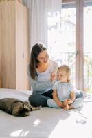 sonriente madre peinada pequeño niña pelo con un amplio cepillo mientras sentado en el cama con su foto