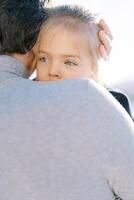 Little girl peeks out from behind her daddy shoulder while sitting in his arms. Back view photo