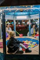 Little girl catches fish with a toy fishing rod while sitting by a toy pool at a fair with children. Back view photo