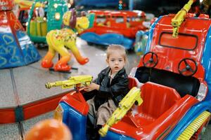 pequeño niña se sienta en un juguete coche en un carrusel, participación el direccion pistola de ruedas foto