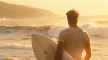 een Mens staand Aan een zanderig strand, Holding een surfboard onder zijn arm Aan een zonnig dag. video