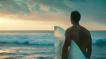 uma homem carrinhos em uma praia, segurando uma prancha de surfe debaixo dele braço. video