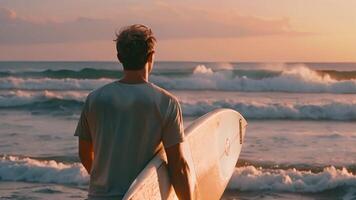 uma homem em pé em a arenoso praia, agarrando uma vibrante prancha de surfe debaixo dele braço, pronto para uma dia do equitação ondas dentro a oceano. video