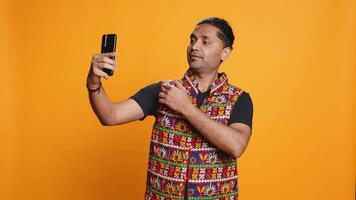 indiano narcisistica uomo utilizzando cellulare per prendere selfie a partire dal tutti angoli. vanitoso sociale media utente assunzione fotografie utilizzando Telefono autoscatto telecamera, sorridente felicemente, studio sfondo, telecamera B video