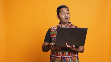 Indian man having entertaining conversation with friends during teleconference meeting using laptop, studio background. Happy person interacts with mates during online videocall on notebook, camera B video