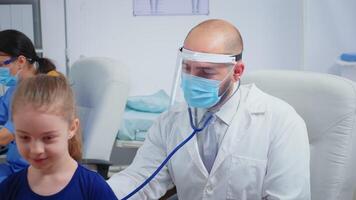 Male doctor giving girl checkup in doctor office using stethoscope during coronavirus. Physician specialist in medicine providing health care services consultation treatment examination in hospital video