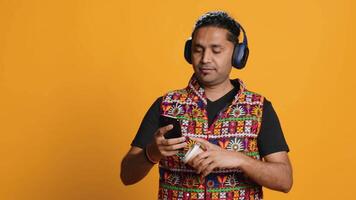 Joyous man listening music, having fun dancing, enjoying hot beverage. Upbeat person wearing headphones, doing funky movements, drinking coffee, studio background, camera A video