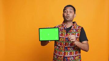 Upset indian man shows thumbs down sign gesturing holding isolated screen tablet, disapproving. Person does rejection hand gesture regarding mockup device, studio background, camera A video