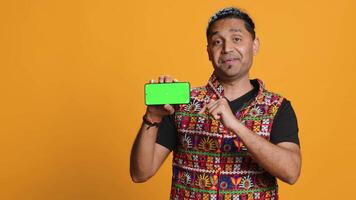 Smiling indian man presenting cellphone with green screen display, isolated over studio background. Person in traditional clothing creating promotion with blank copy space mockup phone, camera A video