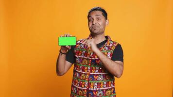 Smiling indian man presenting cellphone with green screen display, isolated over studio background. Person in traditional clothing creating promotion with blank copy space mockup phone, camera B video