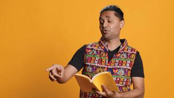 Joyous indian teacher reading book for pupils during class, isolated over studio background. Cheerful man holding novel, telling story to kindergarten kids, teaching them, camera A video