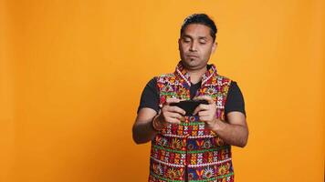 Cheerful Indian person in traditional clothing entertained by videogames on smartphone. Joyous player enjoying game on phone, having fun online, studio backdrop, camera B video