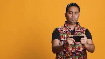 glücklich indisch Mann unterhalten durch Videospiele auf Smartphone, genießen Freizeit Zeit. Spieler genießen Spiel auf Telefon, haben Spaß besiegen Feinde, isoliert Über Studio Hintergrund, Kamera ein video