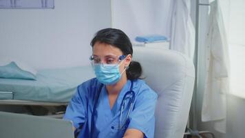 Ward assistant having protection mask and glasses writing a report on laptop. Physician specialist in medicine providing health care services consultation treatment in hospital cabinet during covid-19 video