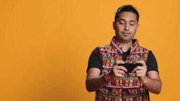Cheerful Indian person in traditional clothing entertained by videogames on smartphone. Joyous player enjoying game on phone, having fun online, studio backdrop, camera A video