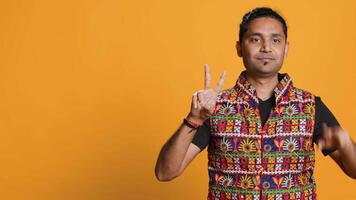 Portrait of upbeat man crossing fingers to make wish, hoping for good luck, isolated over studio background. Jolly indian person doing peace victory hand sign gesture, camera A video