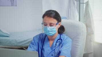 Paramedic with protection mask sitting in doctor office writing on laptop during coronavirus. Physician specialist in medicine providing health care services consultation treatment in hospital cabinet video