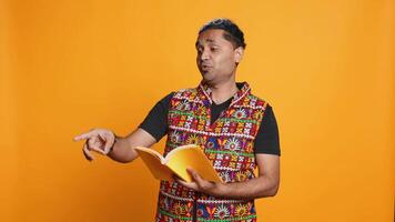 Joyous indian teacher reading book for pupils during class, isolated over studio background. Cheerful man holding novel, telling story to kindergarten kids, teaching them, camera B video