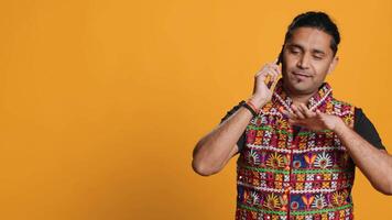 Happy man in traditional clothing chatting with best friend over telephone call. Upbeat person with colorful attire communicating with mate using phone, studio background, camera B video
