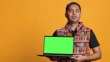Upbeat tech specialist doing promotion using isolated screen laptop, studio background. Cheerful indian person holding chroma key notebook, doing advertisement for it, camera A video