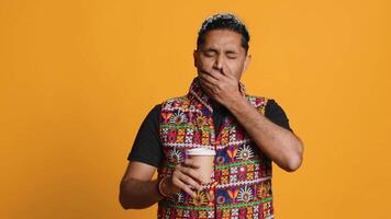 Tired indian man sipping fresh coffee from disposable paper cup early in the morning to wake up and be energized. Person drinking hot beverage from recycled takeaway cup, studio background, camera A video