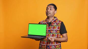 Tech content creator doing influencer marketing using green screen laptop, isolated over studio background. Indian man holding empty copy space mockup notebook, doing review, camera B video