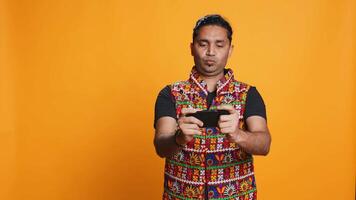 glücklich indisch Mann unterhalten durch Videospiele auf Smartphone, genießen Freizeit Zeit. Spieler genießen Spiel auf Telefon, haben Spaß besiegen Feinde, isoliert Über Studio Hintergrund, Kamera b video