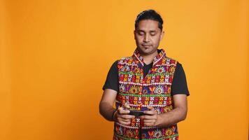 Indian man playing videogames on cellphone in landscape mode, enjoying leisure time. Gamer enjoying game on mobile phone, having fun, isolated over studio background, camera B video