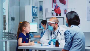 Little girl speaking with doctor during consultation for covid-19. Pediatrician specialist in medicine with protection mask providing health care services treatment examination in hospital cabinet video