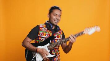 Happy man holding electric guitar, playing rock music, isolated over studio background. Cheerful indian musician using musical instrument, doing concert, playing songs, camera B video
