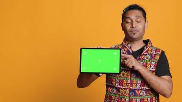 Portrait of indian man doing influencer marketing using green screen tablet, isolated over studio background. Smiling person holding empty copy space mockup device, camera A video